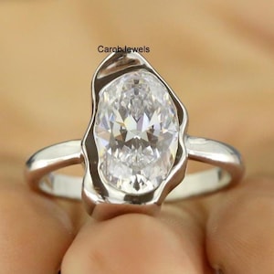 May include: A silver ring featuring a large, oval-cut, clear gemstone. The gemstone is set in a unique, organic-shaped bezel. The ring's band is a simple, polished silver. The brand name "CarobJewels" is visible.