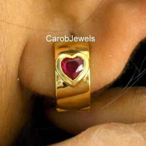 May include: A gold hoop earring featuring a heart-shaped, ruby-red gemstone. The earring is worn on an ear, with the brand name "CarobJewels" visible. The earring is a simple, elegant design.