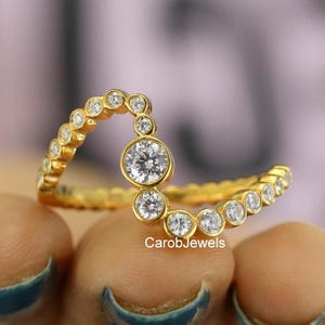 May include: A gold ring featuring a curved, wave-like band set with a series of small, round, clear stones in bezel settings. The center of the curve is accented by a slightly larger round clear stone. The text CarobJewels is visible on the band.