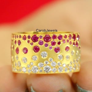 May include: A wide gold ring with a textured surface, adorned with a pattern of red and white gemstones. The ring is held in a hand against a red background. The brand name "CarobJewels" is visible.
