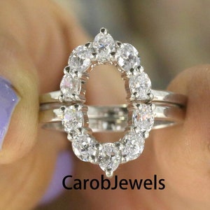 Puede incluir: Un anillo de plata con un halo de pequeños diamantes, formando una forma ovalada. El anillo está puesto en un dedo con esmalte de uñas morado. El texto "CarobJewels" es visible debajo del anillo.