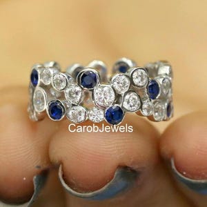 Può includere: Un anello d'argento dal design unico, con pietre preziose rotonde, chiare e blu scuro. L'anello è incastonato con più pietre, creando un aspetto strutturato e a bolle. Il nome del marchio "CarobJewels" è visibile.