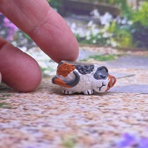 Może przedstawiać: Miniaturowa figurka kota trójkolorowego zwiniętego w kłębek. Kot ma białe umaszczenie z pomarańczowymi i ciemnoszarymi łatami. Figurka jest na tyle mała, że mieści się między palcami i posiada wyraźnie zaznaczone detale pyszczka.