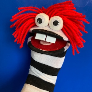 May include: A handmade sock puppet with red yarn hair, large googly eyes, and two white square teeth. The puppet's mouth is red with a black border, and the sock has black and white stripes.