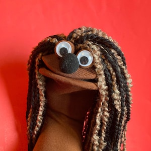 May include: A brown sock puppet featuring a round black nose and two large googly eyes. The puppet has long, thick yarn hair in alternating strands of black and tan, which hangs down on both sides of its face. The mouth is open, revealing a brown interior.