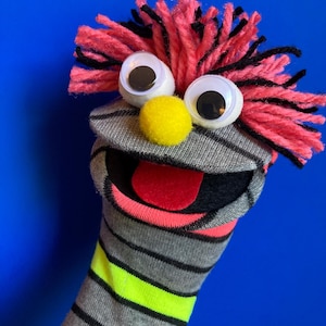 May include: A sock puppet featuring a grey and black striped body with a neon yellow band. It has a bright yellow pom-pom nose, two large googly eyes, and a red felt tongue inside a black mouth. The hair is made of messy pink and black yarn.