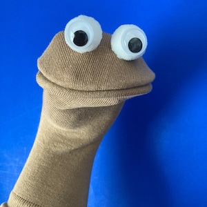 May include: A tan sock puppet with large, googly eyes against a bright blue background. The sock is made of a ribbed knit material. The eyes are white with black pupils, giving the puppet a whimsical appearance. The puppet's mouth is slightly open.