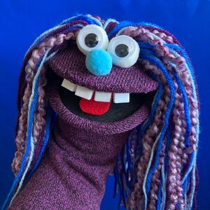 May include: A purple sock puppet featuring long, multi-colored yarn hair in shades of blue, purple, and pink. The puppet has large googly eyes, a bright blue pom-pom nose, white square teeth, and a red felt tongue inside an open mouth.