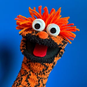 May include: A hand puppet made of orange and black textured fabric with bright orange yarn hair. It features two large white googly eyes, a small orange pom-pom nose, and a wide open mouth with a red felt tongue visible inside the black interior.