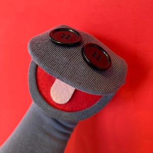May include: A handmade sock puppet with a gray body, red mouth, and a white felt tongue. Two large black buttons serve as eyes. The puppet is set against a vibrant red background.