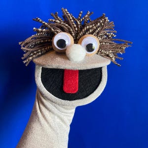 May include: A beige sock puppet featuring a mop of brown and white yarn hair, two large googly eyes, a white pom-pom nose, and a wide open mouth with a bright red felt tongue. The puppet is constructed from a light tan fabric sock base.