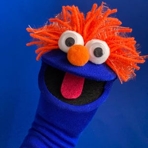 May include: A blue sock puppet with orange yarn hair, large googly eyes, and an orange nose. The puppet's mouth is open, revealing a pink tongue. The background is a solid blue.