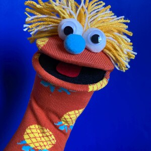 May include: A handmade sock puppet with googly eyes, a blue nose, and yellow yarn hair. The orange sock features a repeating pineapple pattern. The puppet's mouth is open, revealing a red tongue, and the background is a solid blue.