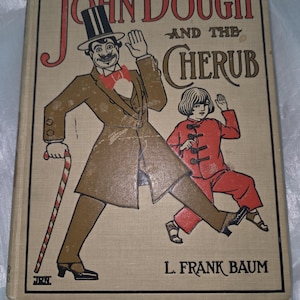May include: A vintage hardcover book titled John Dough and the Cherub by L. Frank Baum. The tan cover features a central illustration of a man in a top hat and brown coat holding a candy cane walking beside a child in a red outfit with black toggles.