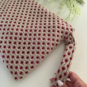 以下が含まれることがあります： ベージュとバーガンディのクラッチバッグ。幾何学的な透かし模様のデザイン。バッグにはお揃いのハンドルが付いており、柔らかく質感のある素材で作られています。内側は濃いバーガンディ色で、手作りのようです。