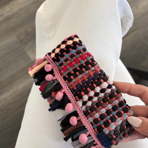 Puede incluir: Un clutch rectangular tejido con un patrón colorido de hilos negros, rojos, azules y grises. El clutch está adornado con pompones rosas y un borde rosa. La franja en la parte inferior incluye varios retazos de tela.
