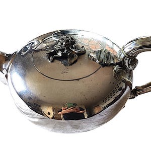 Op de afbeelding: Een zilveren theepot met een gebogen tuit en handvat. Het deksel heeft bloemen- en bladvormige versieringen. De theepot heeft een reflecterend oppervlak en het ontwerp is sierlijk en elegant.