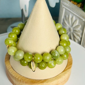 Könnte beinhalten: Ein Armband mit großen, runden, hellgrünen Perlen. Das Armband wird auf einem beigefarbenen, kegelförmigen Schmuckständer mit Holzsockel präsentiert. Ein kleiner goldfarbener Anhänger mit dem Text "1988 ES" hängt am Armband.