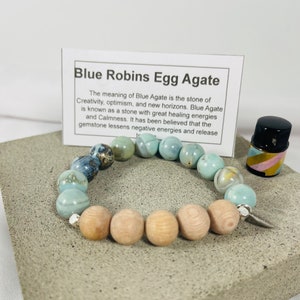 May include: A blue agate gemstone bracelet with wooden beads and a silver charm. The bracelet is on a light grey surface with a small bottle of essential oil. The text on the card reads: "Blue Robins Egg Agate. The meaning of Blue Agate is the stone of Creativity, optimism, and new horizons. Blue Agate is known as a stone with great healing energies and Calmness. It has been believed that the gemstone lessens negative energies and release"