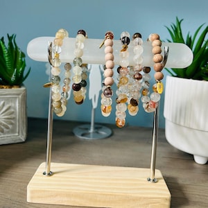 Puede incluir: Un expositor con varias pulseras de cuentas. Las pulseras presentan cuentas transparentes, marrones y de madera. El expositor tiene una base de madera clara y soportes de metal plateado, con una barra horizontal blanca para sujetar las pulseras. La base mide unos 20 cm.