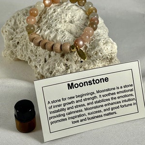 May include: A moonstone bracelet with wooden beads and a gold charm. The bracelet is on a white rock. A small bottle of essential oil is in the lower left corner of the image. The text on the card reads: "Moonstone A stone for new beginnings, Moonstone is a stone of inner growth and strength. It soothes emotional instability and stress, and stabilises the emotions, providing calmness. Moonstone enhances intuition, promotes inspiration, success, and good fortune in love and business matters."