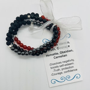 May include: Three layered bracelets made with black lava rock, black obsidian, hematite, and carnelian beads. The bracelets are tied with a white ribbon and a tag that reads "Confidence, Truth, Courage. Hematite, Obsidian, Carnelian. -Dissolves negativity, boosts self-esteem. -Truth, protection. -Courage, confidence."
