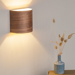 Puede incluir: Una l&aacute;mpara de pared de madera con forma curva y luz blanca c&aacute;lida. La l&aacute;mpara est&aacute; montada en una pared blanca y proyecta un suave resplandor en la pared.