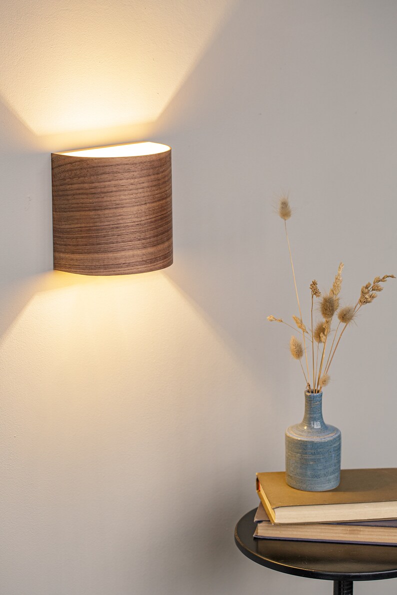 Puede incluir: Una l&aacute;mpara de pared de madera con forma curva y luz blanca c&aacute;lida. La l&aacute;mpara est&aacute; montada en una pared blanca y proyecta un suave resplandor en la pared.