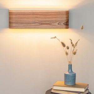 May include: A wooden wall sconce with a warm white light illuminates a white wall. A blue vase with dried flowers sits on a black side table with a stack of books.