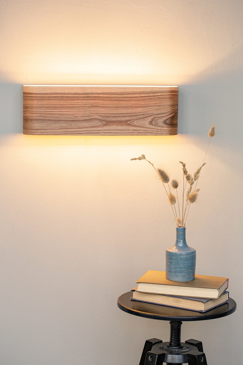 May include: A wooden wall sconce with a warm white light illuminates a white wall. A blue vase with dried flowers sits on a black side table with a stack of books.