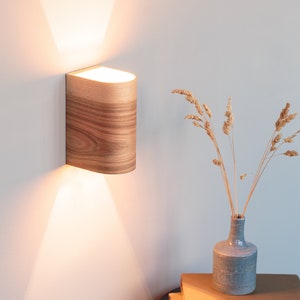May include: A wooden wall sconce with a warm white light casts a beam of light on a white wall. The sconce is mounted on a white wall and has a natural wood grain finish. A vase with dried flowers sits on a black side table with a stack of books.