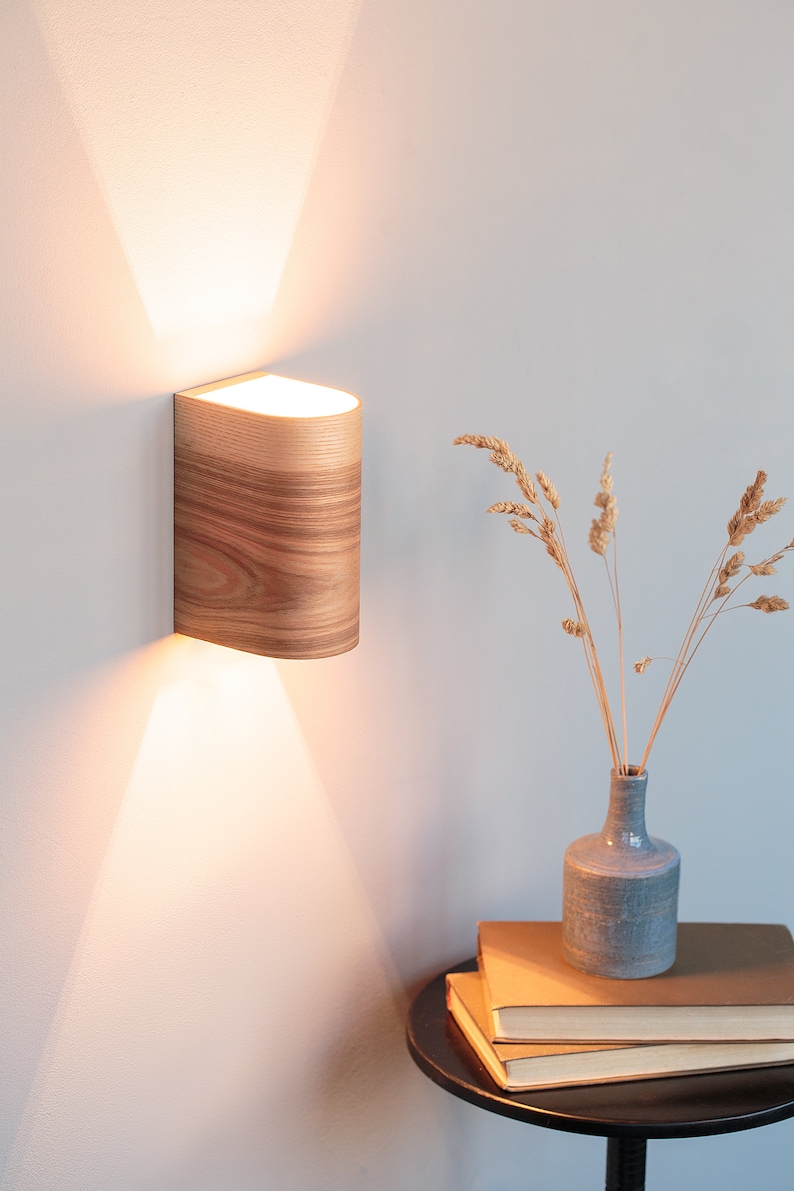 May include: A wooden wall sconce with a warm white light casts a beam of light on a white wall. The sconce is mounted on a white wall and has a natural wood grain finish. A vase with dried flowers sits on a black side table with a stack of books.
