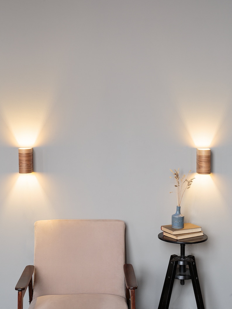 May include: A beige armchair with wooden arms sits in front of a light gray wall with two wooden wall sconces. A small black side table with a vase of dried flowers and a stack of books sits to the right of the chair.