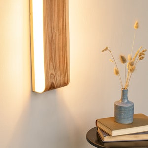 May include: A wooden wall sconce with a curved design and a white light panel. The sconce is mounted on a white wall and casts a warm glow.