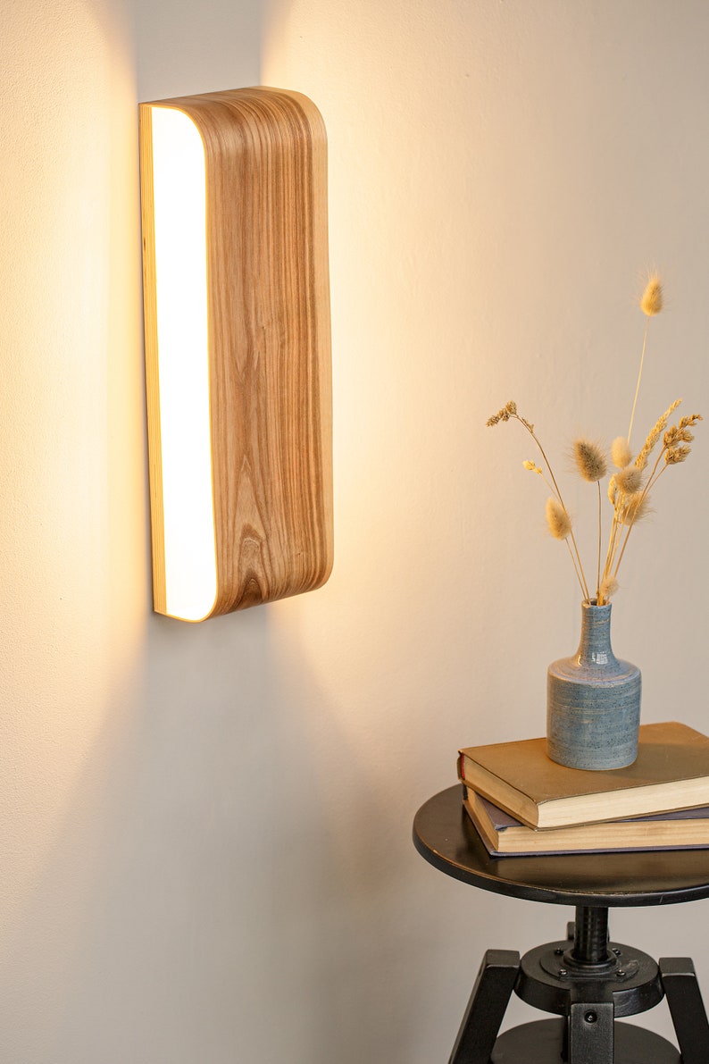 May include: A wooden wall sconce with a curved design and a white light panel. The sconce is mounted on a white wall and casts a warm glow.