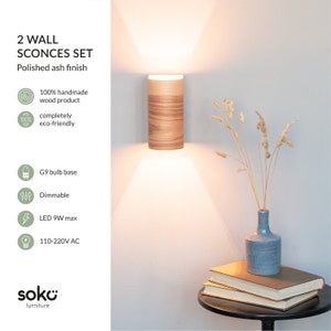 May include: Two polished ash wall sconces with a wood grain finish. The sconces are lit and casting a warm glow. The sconces are 100% handmade and eco-friendly. They are dimmable and use a G9 bulb base. The sconces are made by soko furniture.