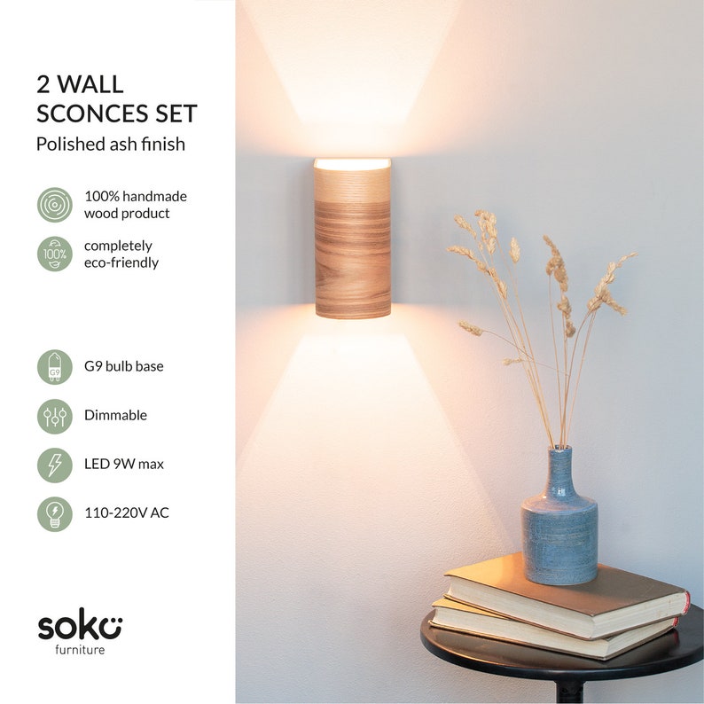 May include: Two polished ash wall sconces with a wood grain finish. The sconces are lit and casting a warm glow. The sconces are 100% handmade and eco-friendly. They are dimmable and use a G9 bulb base. The sconces are made by soko furniture.