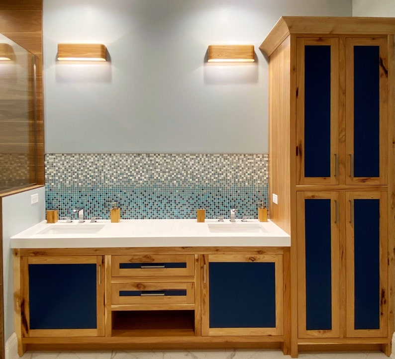 May include: A bathroom vanity with two sinks and a cabinet with blue doors. The vanity is made of wood and has a white countertop. The cabinet has two drawers and a shelf. The bathroom has a tiled wall with a mosaic pattern.