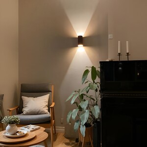 May include: A living room interior with a gray armchair, a black piano, a potted plant, and a wooden coffee table. The wall is painted a light gray color and has a wall sconce with a brown cylindrical shade.