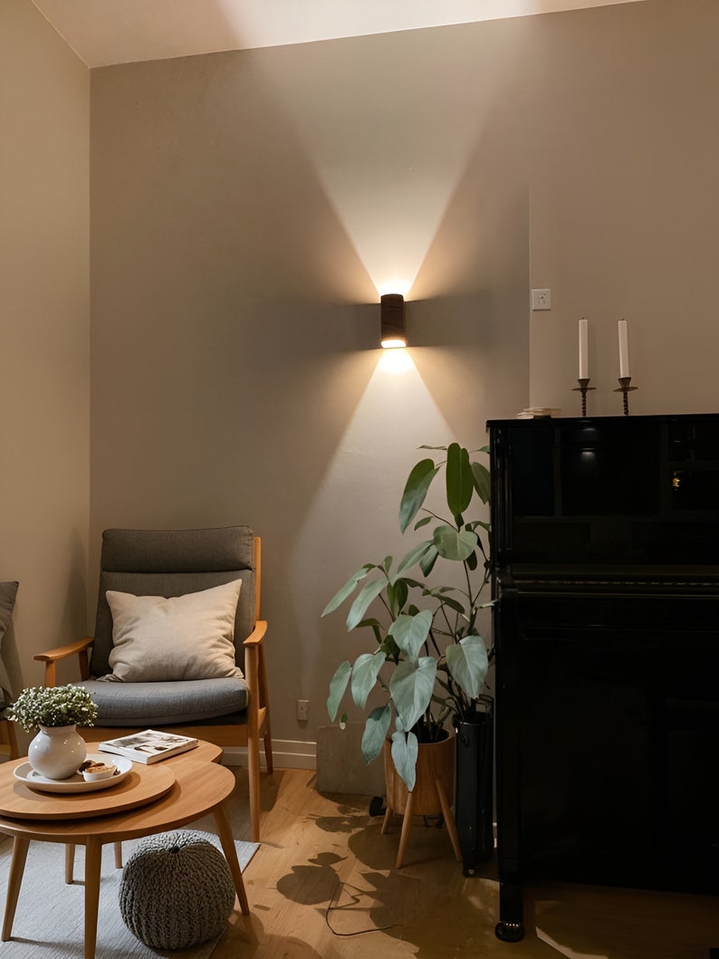 May include: A living room interior with a gray armchair, a black piano, a potted plant, and a wooden coffee table. The wall is painted a light gray color and has a wall sconce with a brown cylindrical shade.