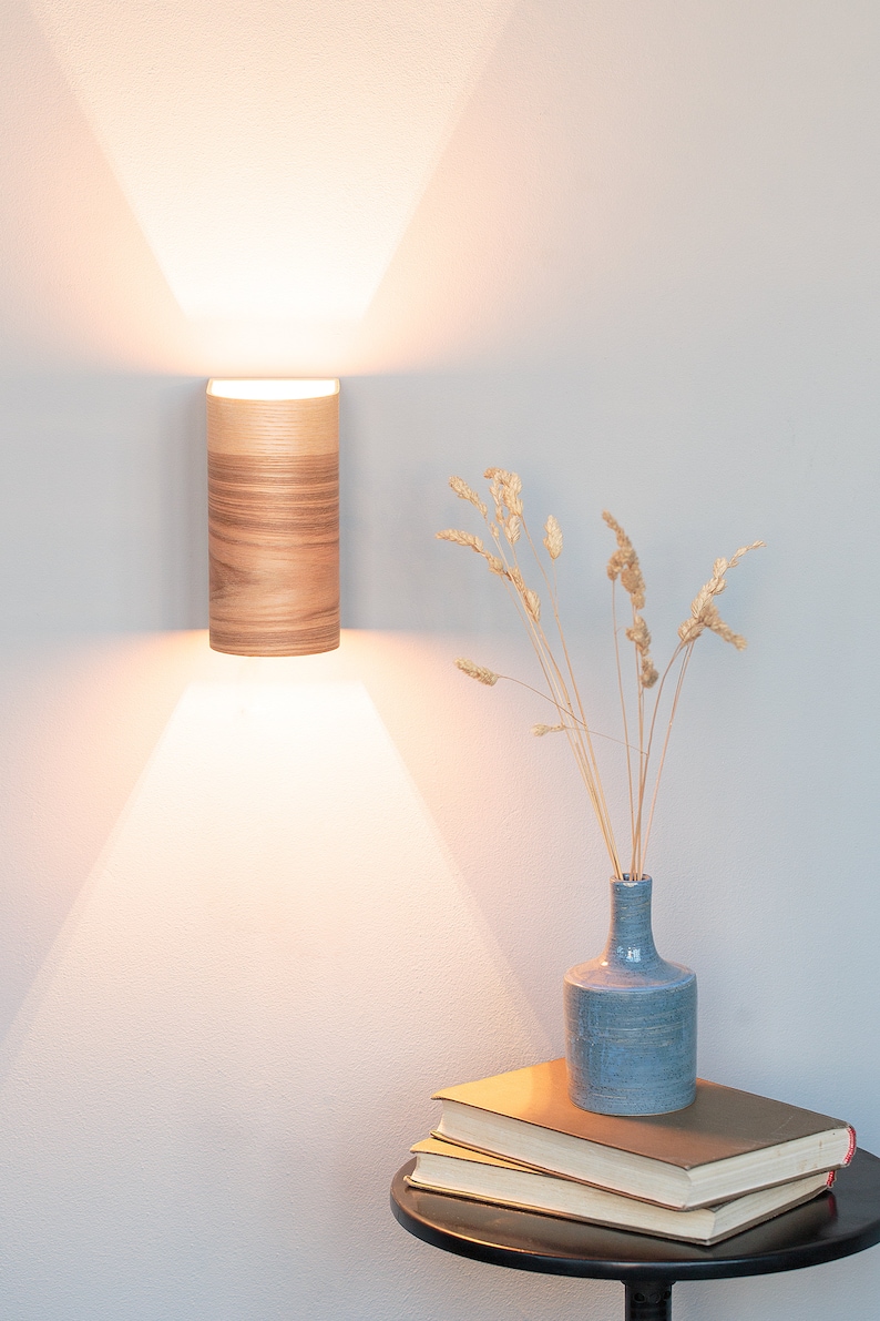 May include: A wooden wall sconce with a warm white light illuminates a small table with a blue vase filled with dried grasses and a stack of books.
