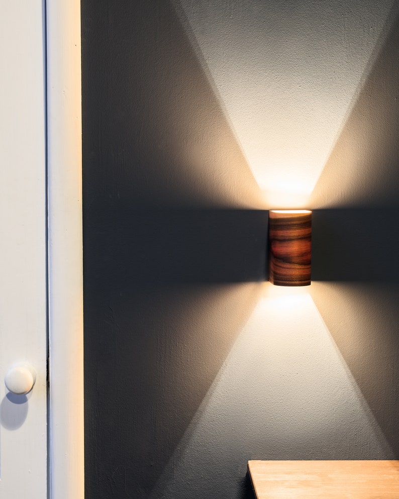 May include: A wooden cylindrical wall sconce with a warm white light shining down on a wooden surface.