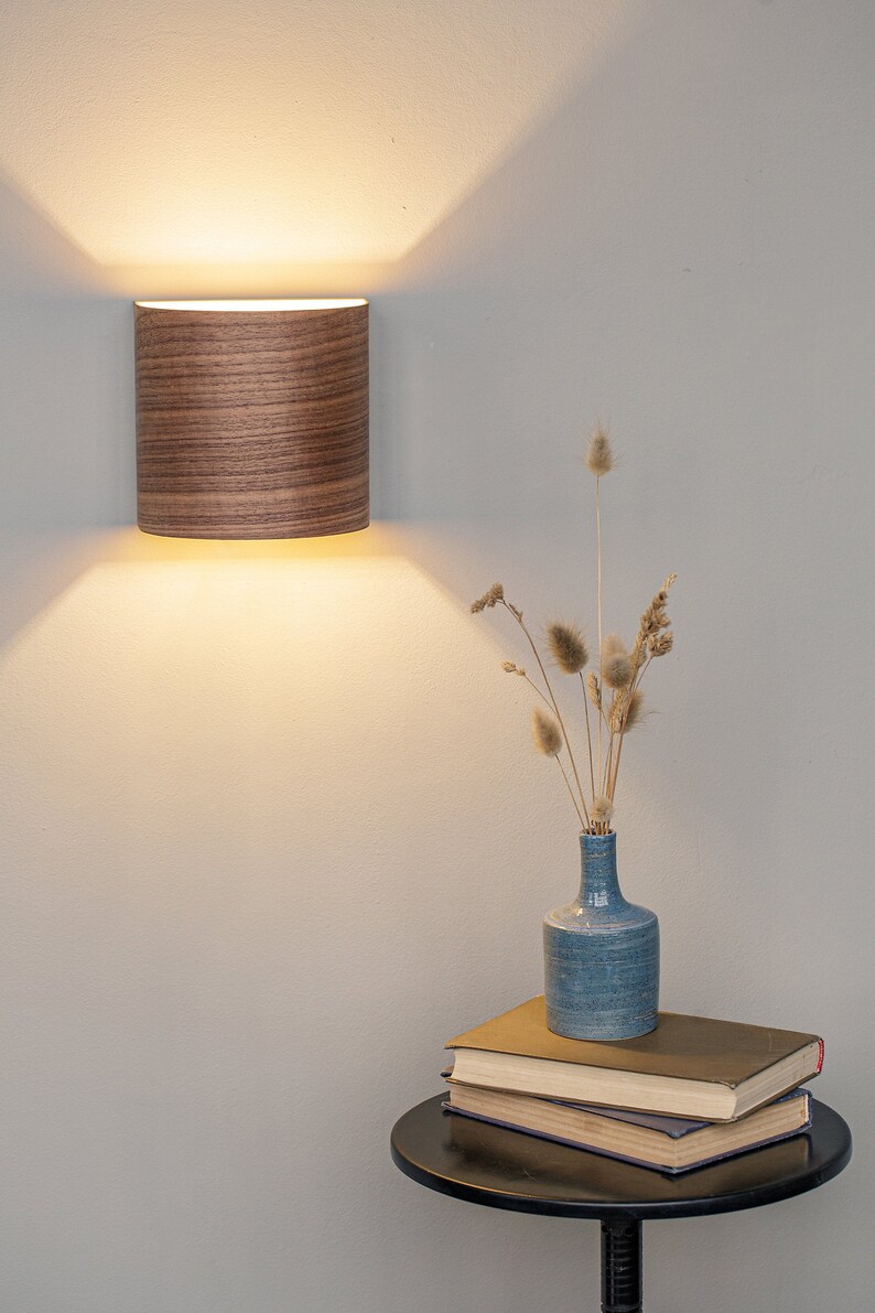 Puede incluir: Una l&aacute;mpara de pared de madera con luz blanca ilumina una pared blanca. Un jarr&oacute;n azul con hierbas secas se encuentra sobre una mesa auxiliar negra con una pila de libros.