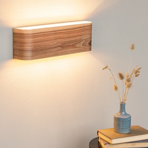 May include: A wooden wall sconce with a warm white light. The sconce is mounted on a white wall and is illuminated, casting a soft glow on the wall.