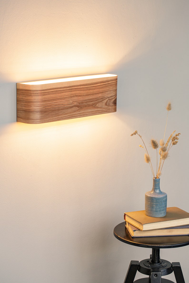 May include: A wooden wall sconce with a warm white light. The sconce is mounted on a white wall and is illuminated, casting a soft glow on the wall.