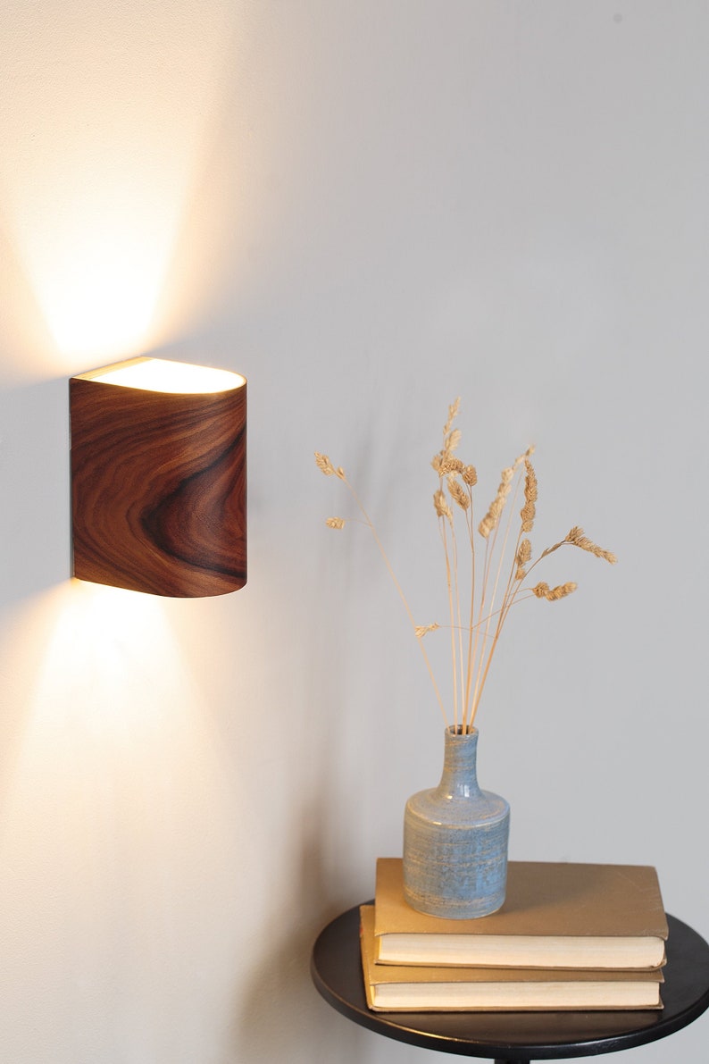 May include: A wooden wall sconce with a curved shape and a light shining from inside. A blue ceramic vase with dried flowers sits on a stack of books on a black side table.