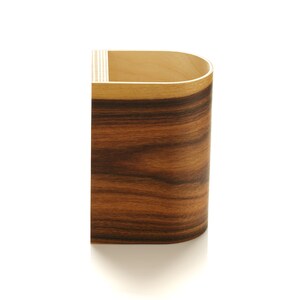 May include: A wooden pencil holder with a rounded top and a dark brown wood grain pattern. The holder is made of wood veneer and has a light brown top edge.