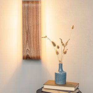 May include: A wooden wall sconce with a warm light illuminates a small table with a blue vase filled with dried grasses and a stack of books.