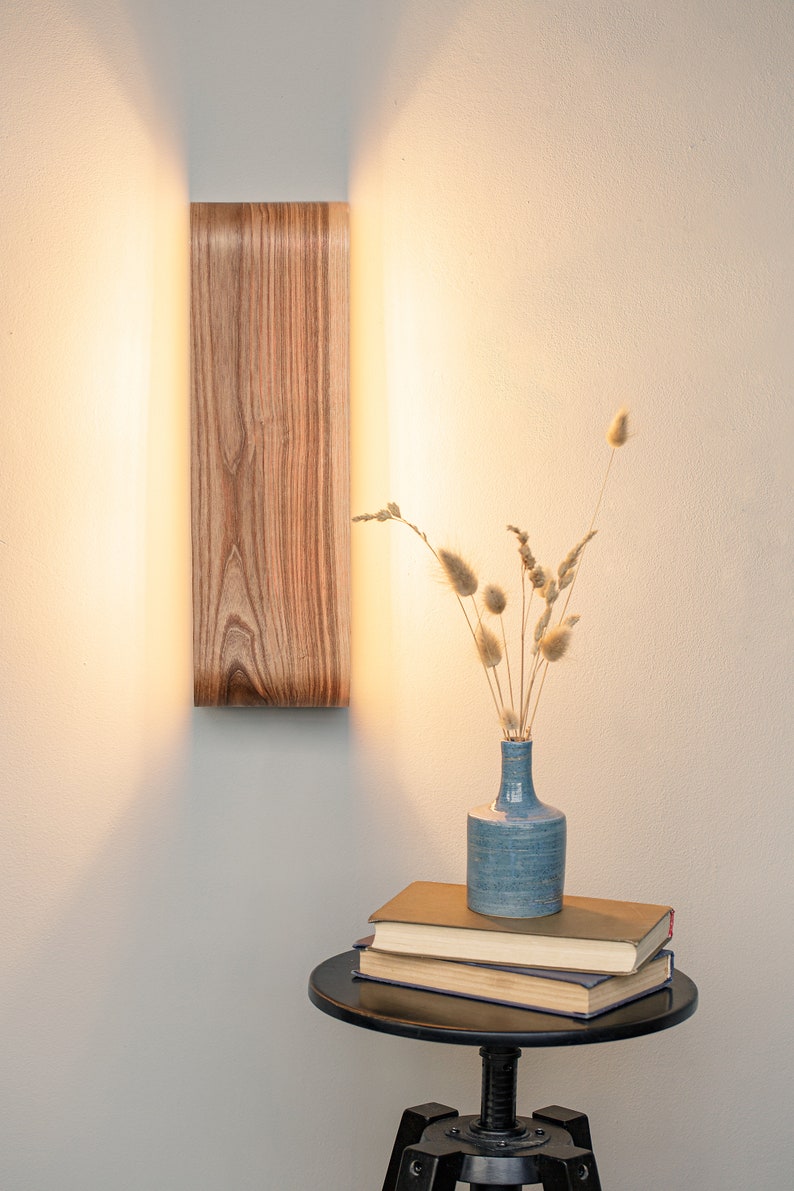 May include: A wooden wall sconce with a warm light illuminates a small table with a blue vase filled with dried grasses and a stack of books.