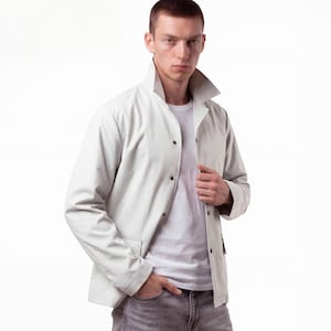 May include: A light gray jacket with a wide collar and black buttons. The jacket is worn over a white t-shirt and paired with gray jeans. The person is standing in front of a white background.